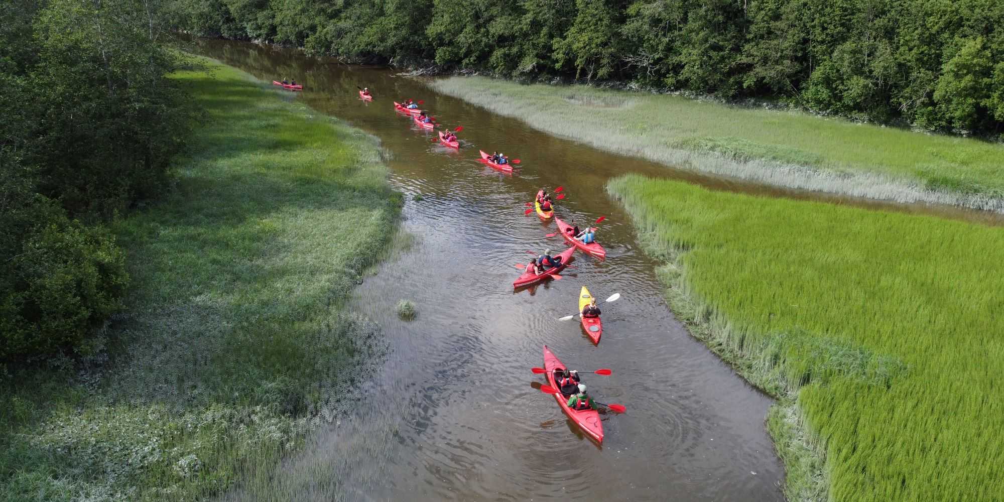 Several red kayaks on a small river wedged between green meadows.